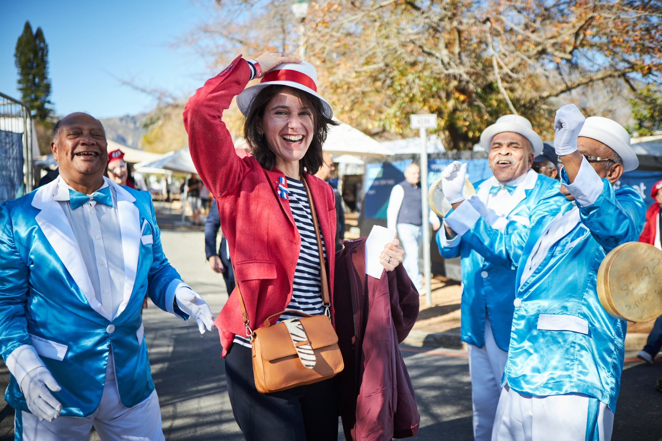 Woman in red jacket and white hat smiles, walking with Kaapse Klopse