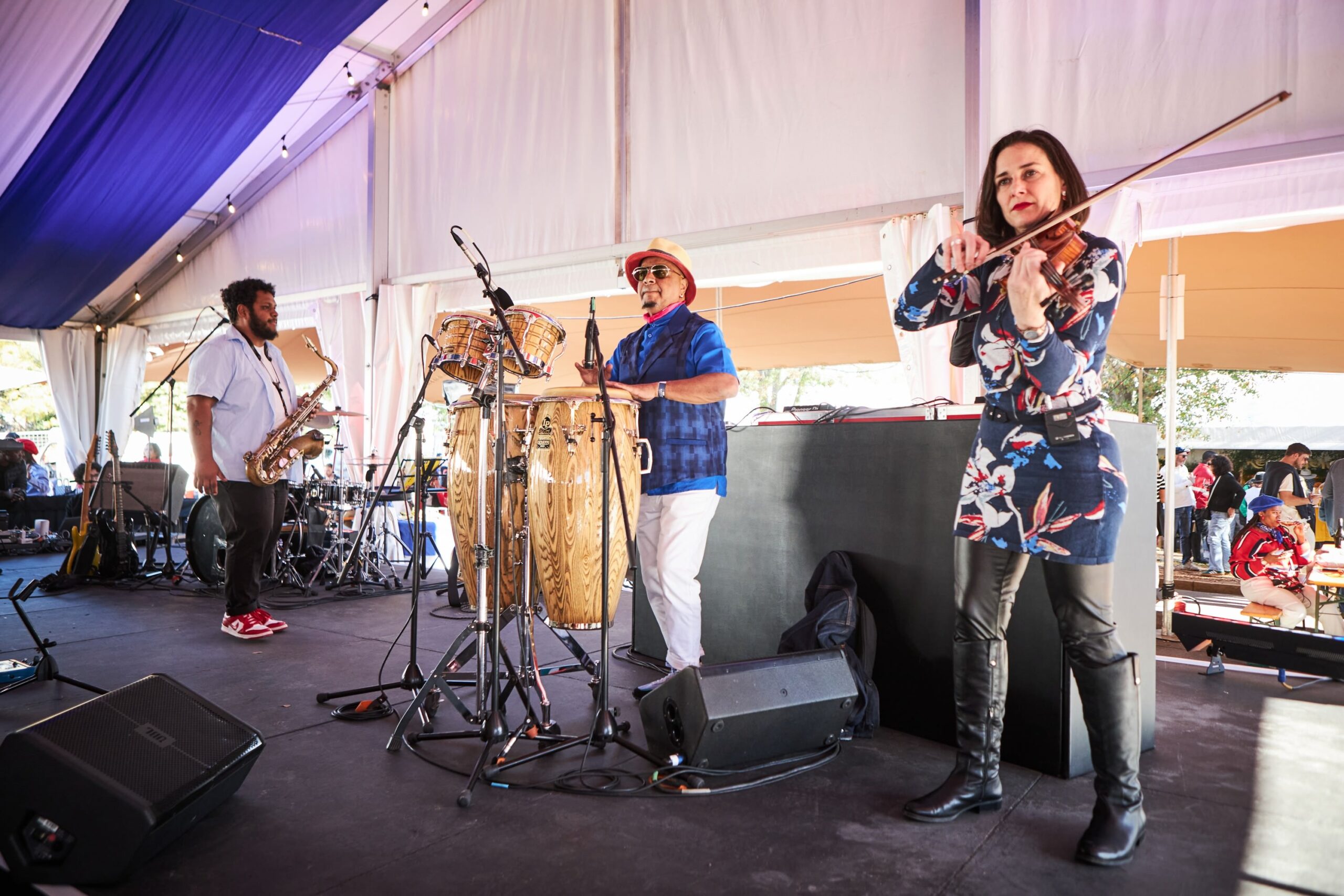 Musicians play music under a tent: saxophonist, conga drummer, and violinist.