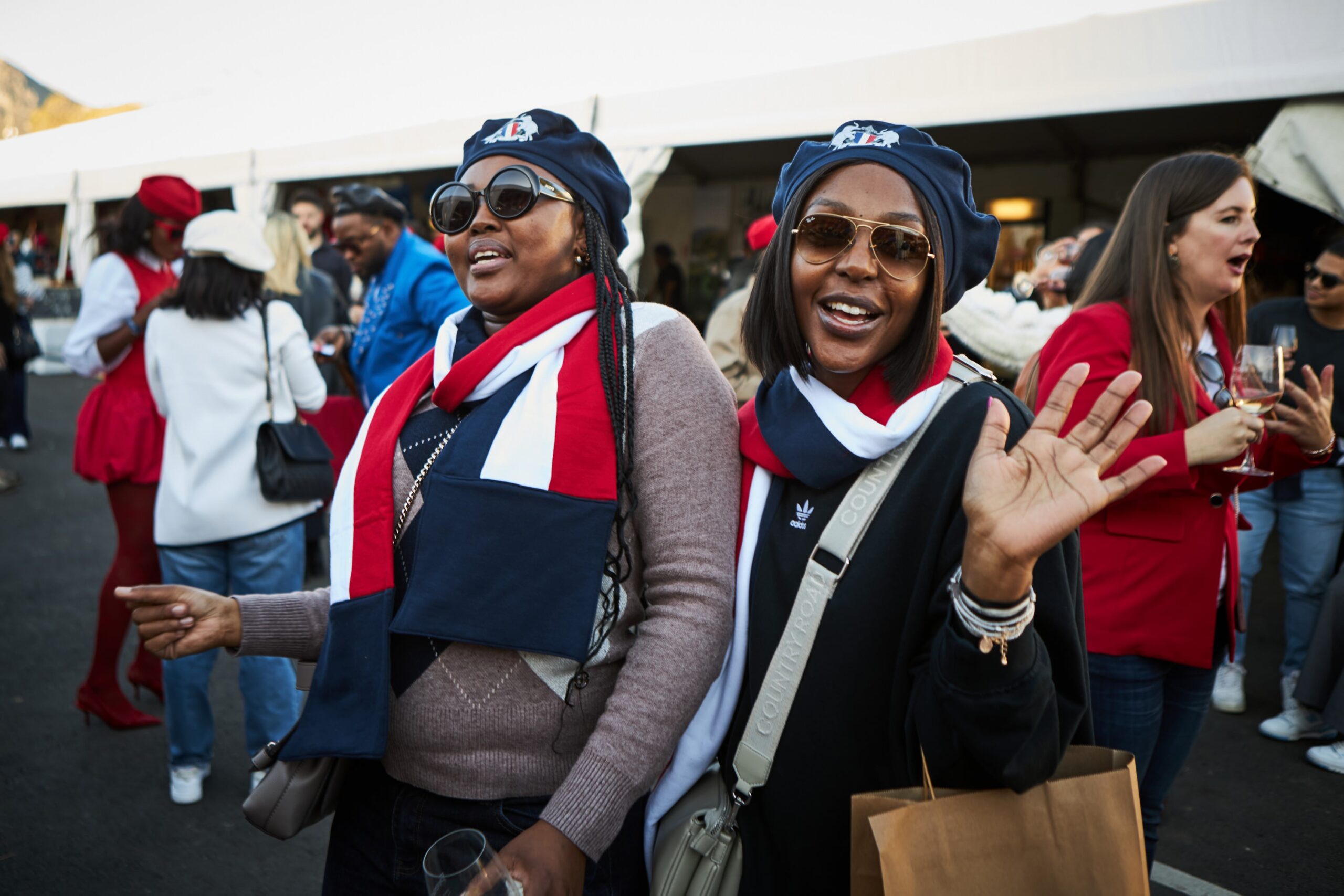 Two women in matching scarves and hats wave at the festival, smiling.